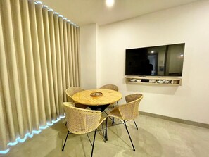 A room with a round wooden table and wicker chairs, a TV mounted on the wall, and a window with blinds. - Luxury Apartment in Santa Marta | Ocean View (Santa Marta)