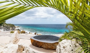 A stone hot tub surrounded by rocks and a wooden deck overlooking a beach with clear turquoise water. - Villa Ocean’s - Luxury Beachfront - Coral Estate (Sint Willibrordus)