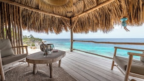A wooden deck with a thatched roof, a table with a champagne bottle and glasses, and a view of the ocean.