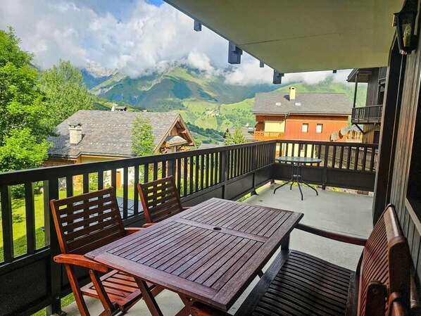 A balcony with wooden furniture, a view of mountains, and a house in the background.