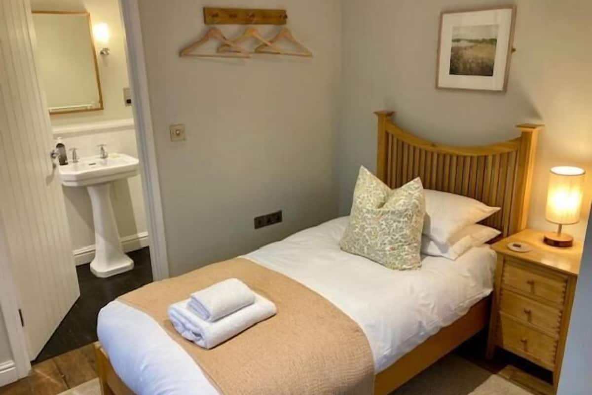 Una camera da letto con un letto, cuscini, un comodino con lampada, una mensola notte, una testiera in legno, un quadro appeso al muro e un bagno con lavandino e specchio.