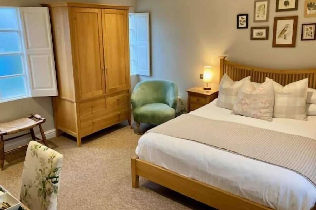 Una camera da letto con un letto, un armadio in legno, una poltrona verde, un tavolino e un comodino con una lampada.