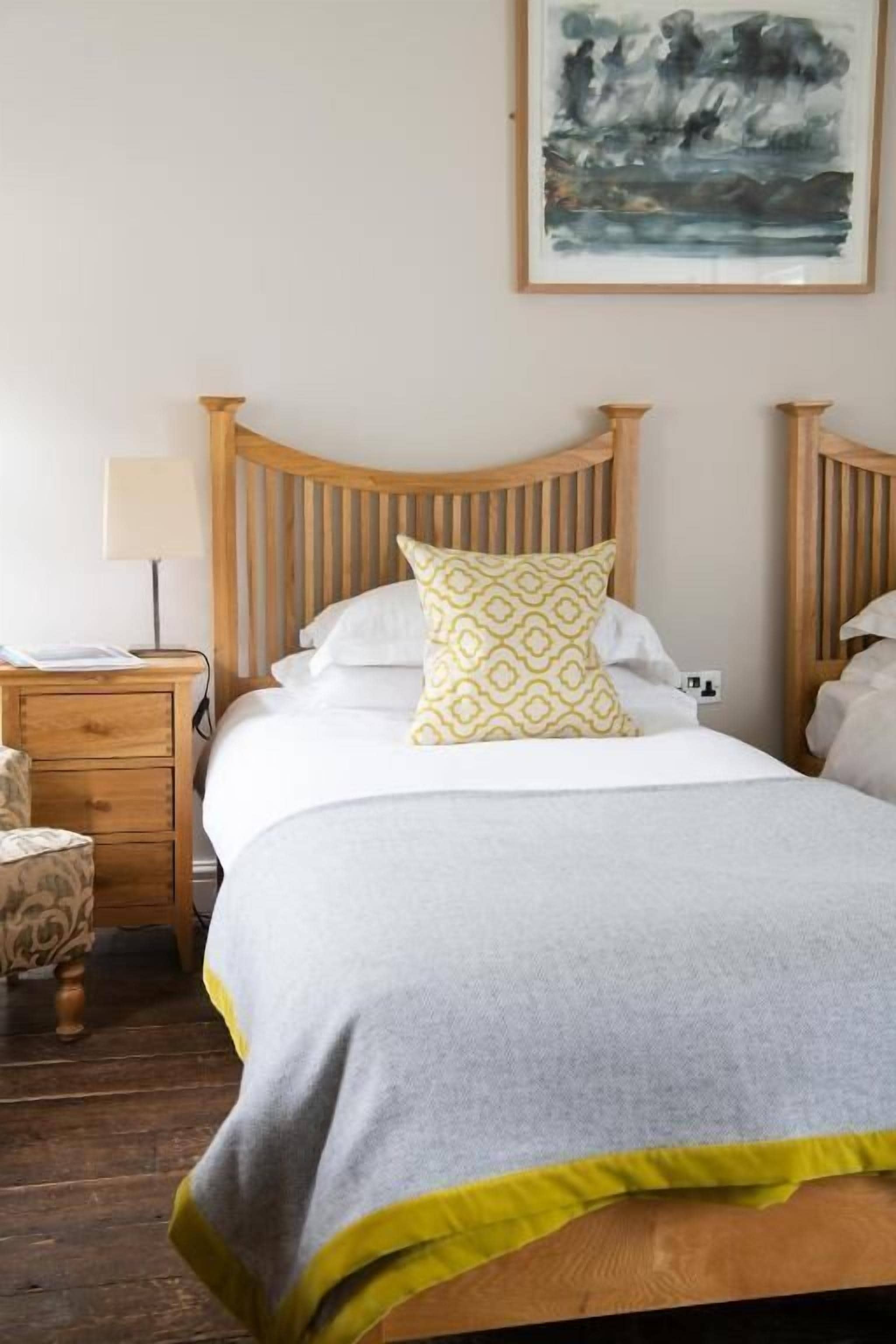 Un dormitorio con cabecera de madera, cama con manta gris y almohada amarilla, mesita de noche con lámpara y un cuadro colgado en la pared.
