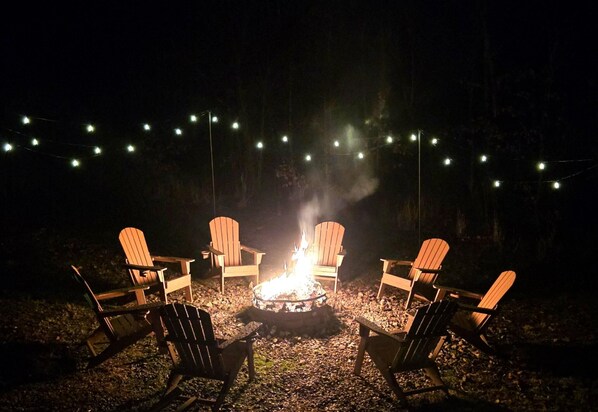 A campfire with chairs around it, string lights overhead, and a forest background.
