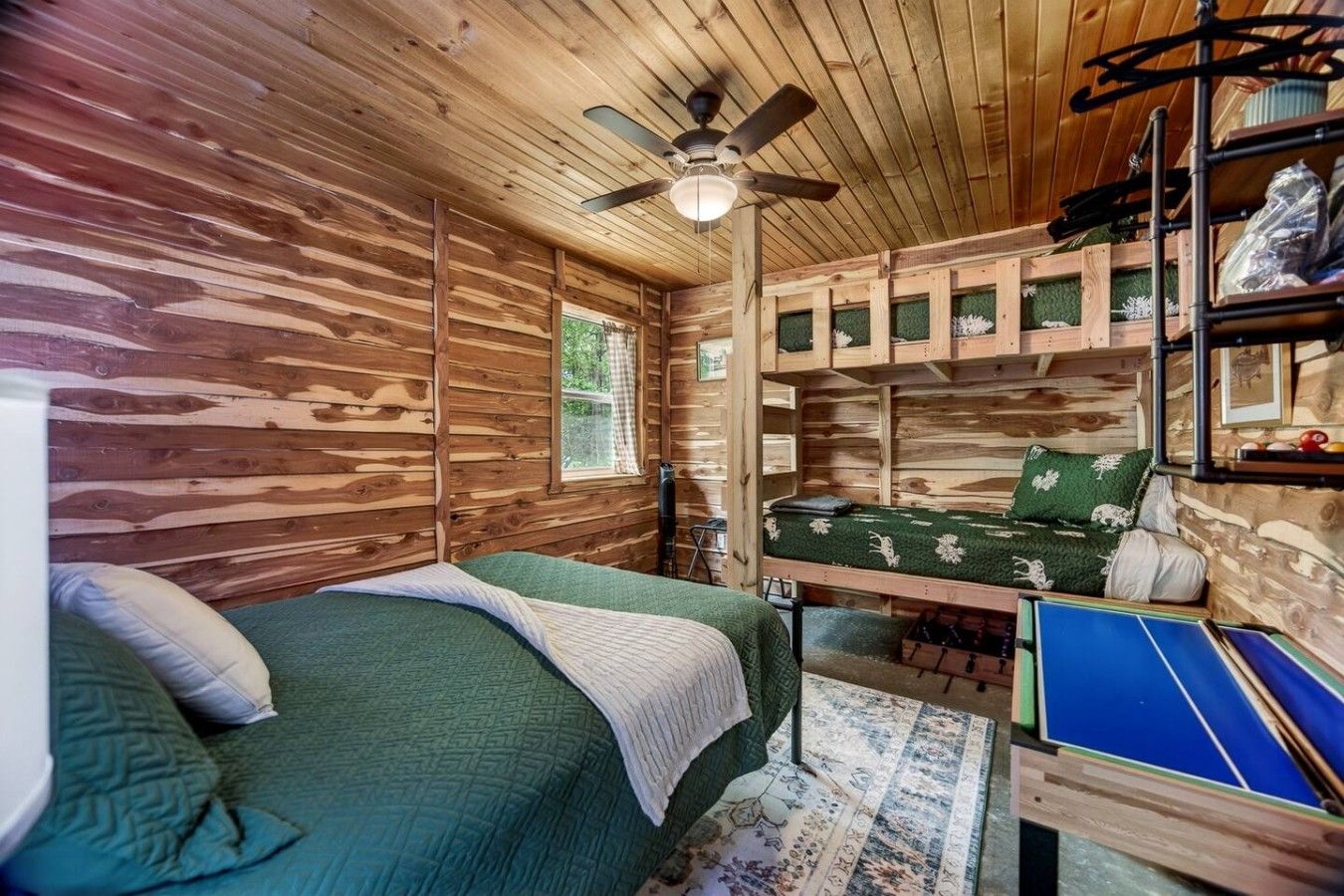 A cabin interior with a bed, a ceiling fan, a small table, and a shelf with items.