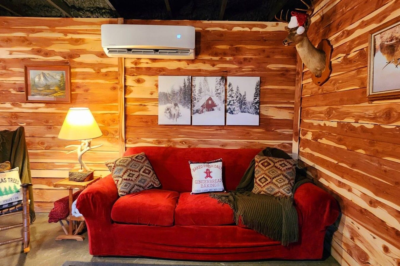 A cozy cabin interior with a red sofa, a mounted deer head, and framed pictures on the wall.