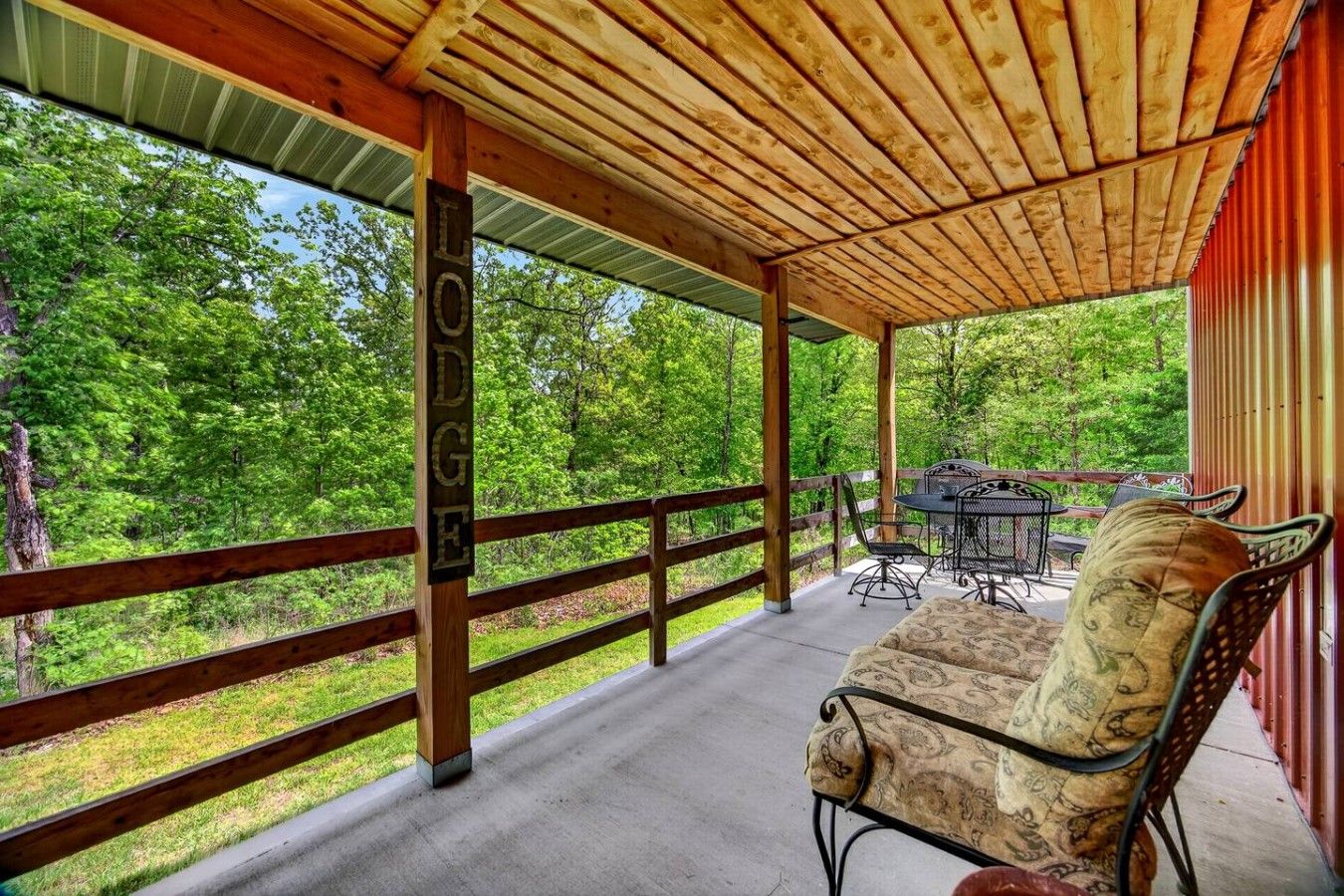 A covered outdoor seating area with a wooden deck, a metal table and chairs, and a view of a forested landscape.