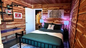 Un dormitori d'estil cabana de fusta amb un llit, làmpades de nit, un escriptori i una porta que dona a una altra habitació.