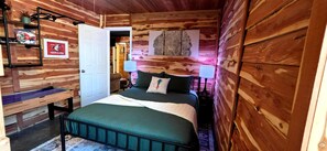 A wooden cabin-style bedroom with a bed, bedside lamps, a desk, and a door leading to another room. - 4 min to Bennett Spring • Dogs Welcome (Lebanon)
