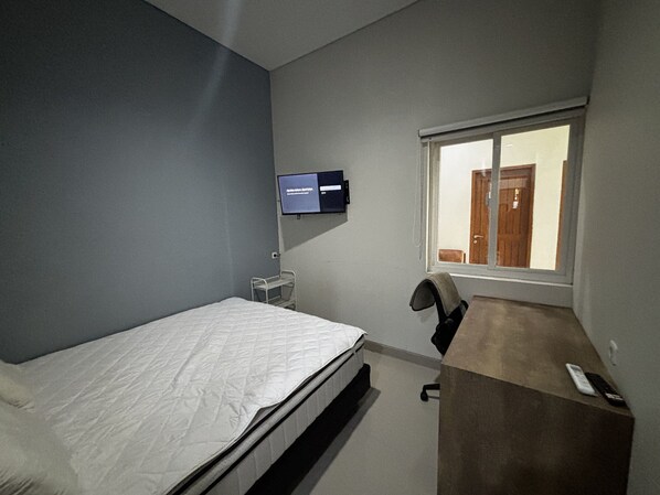 A bedroom with a bed, a desk, a chair, a TV mounted on the wall, and a window with a view of a door.