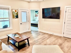 Un salon avec une table basse en bois, un téléviseur fixé au mur et une porte donnant sur une autre pièce.
