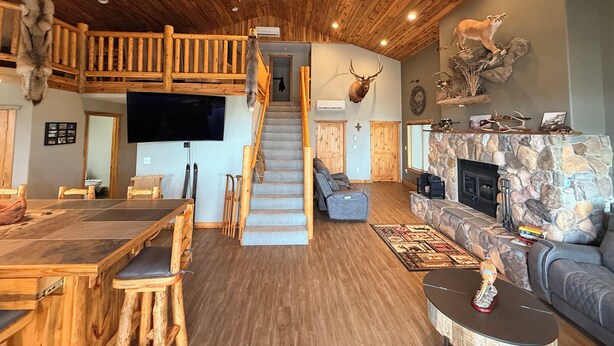 A spacious living area with a wooden ceiling, a stone fireplace, a mounted deer head, and a staircase leading to an upper level. - Spacious 3-bedroom building in Cow creek SD close to Outpost Lodge with AC (Pierre)