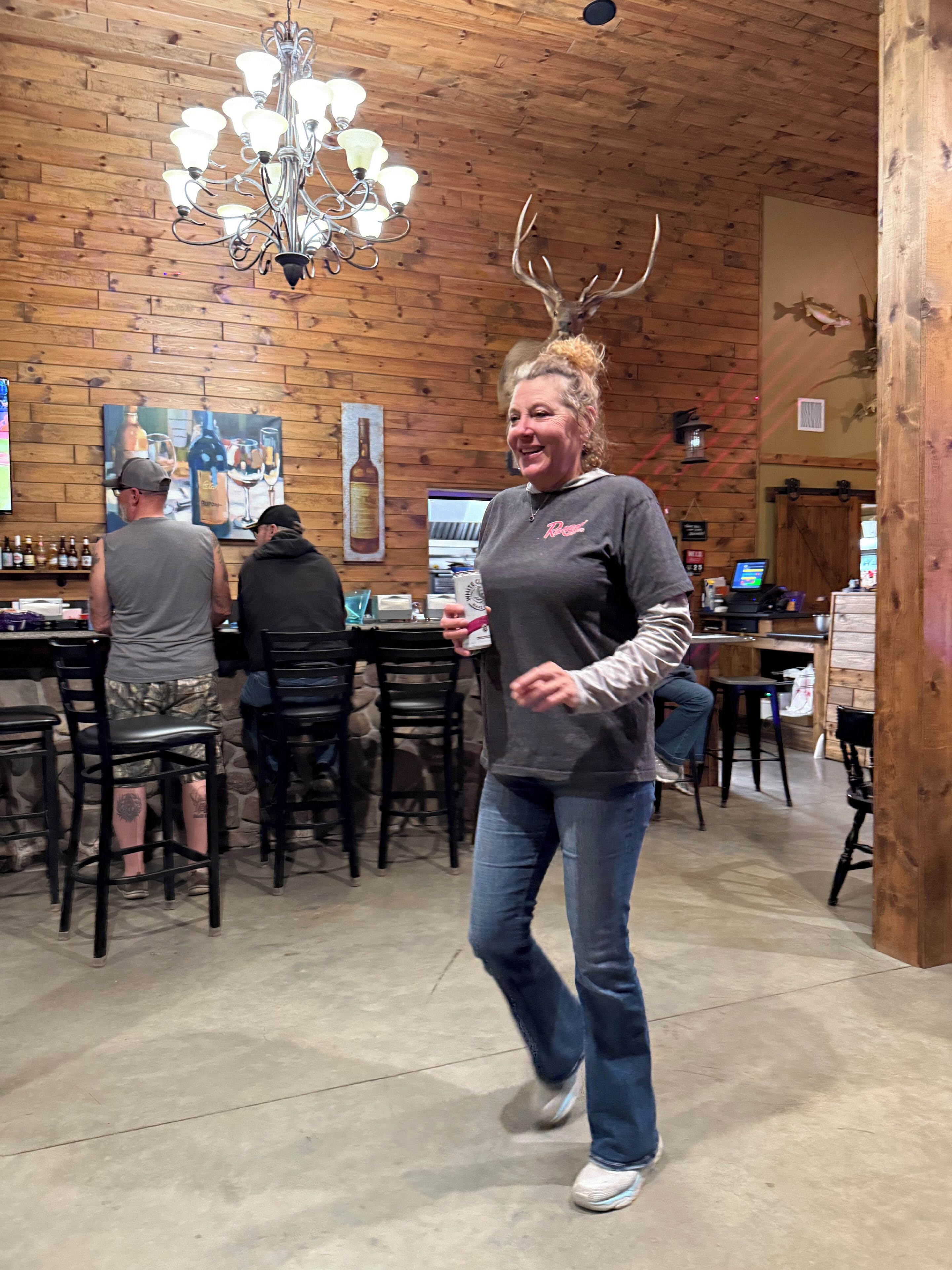 Une personne se promène dans un restaurant aux murs en bois, orné d’un lustre et doté de tables et de chaises.