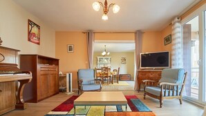 Ett vardagsrum med ett piano, två fåtöljer, en tv och en matplats i bakgrunden.