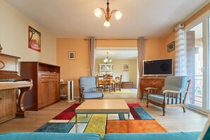 Ein Wohnzimmer mit einem Klavier, zwei Sesseln, einem Fernseher und einem Essbereich im Hintergrund.