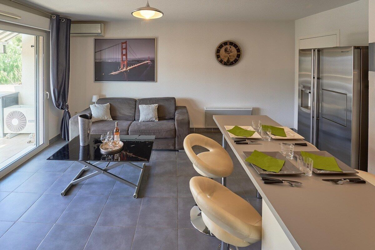 Een moderne woonkamer met een bank, een glazen salontafel, een eethoek met een tafel voor twee personen, een koelkast en een groot raam met gordijnen.