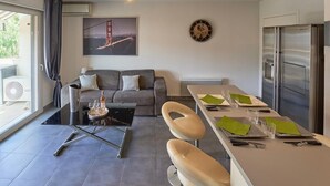 Un salon moderne comprenant un canapé, une table basse en verre, un coin repas avec une table dressée pour deux personnes, un réfrigérateur et une grande fenêtre avec des rideaux.