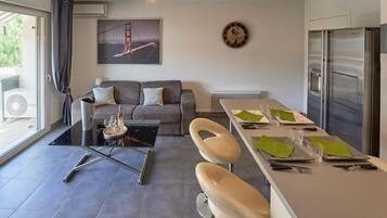 Een moderne woonkamer met een bank, een glazen salontafel, een eethoek met een tafel voor twee personen, een koelkast en een groot raam met gordijnen.