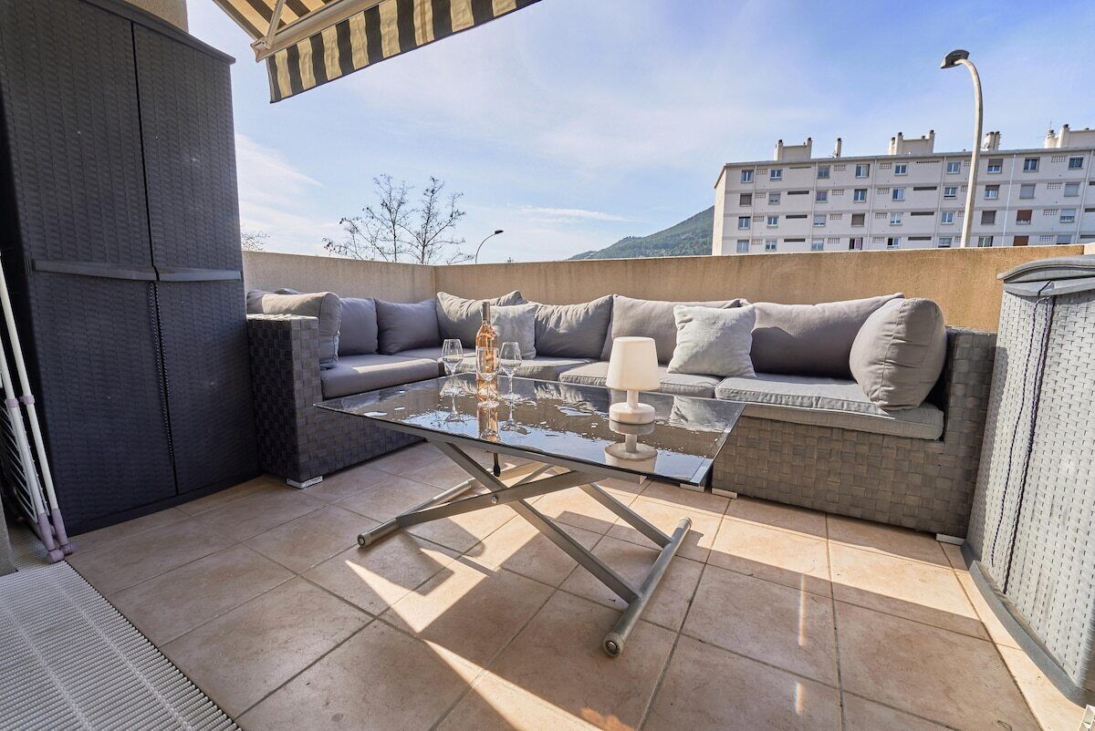 Een terras buiten met een tafel met glazen blad, grijze zitplaatsen met kussens, en een lamp.