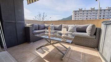 Een terras buiten met een tafel met glazen blad, grijze zitplaatsen met kussens, en een lamp.