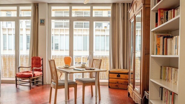 Um quarto com uma grande janela, uma mesa de madeira com cadeiras, uma cadeira vermelha, um armário de madeira e uma estante de livros.