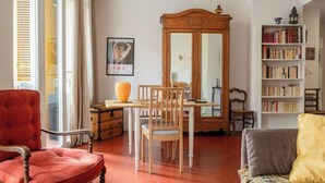 Uma sala de estar com uma mesa de jantar, cadeiras, uma poltrona vermelha, um sofá, uma estante e um espelho grande.