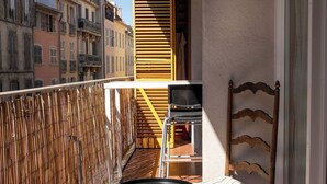 Uma varanda com uma cadeira e uma pequena mesa, uma cadeira de madeira, uma mesa de metal e vista para edifícios.