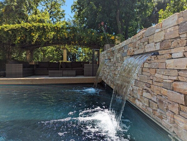 A stone wall with a water feature, a pool, and outdoor furniture.