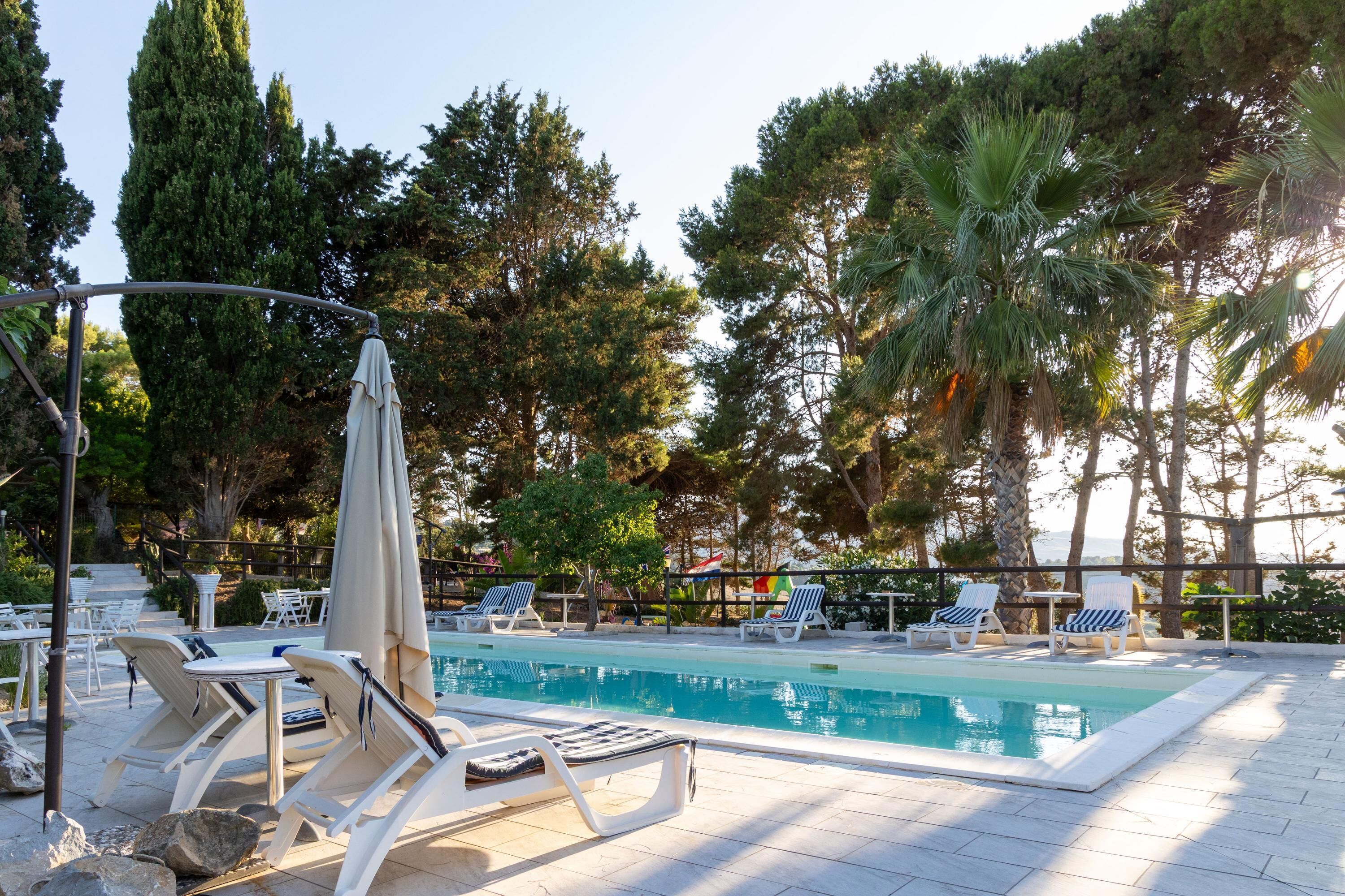 A pool area with lounge chairs and umbrellas, surrounded by trees and a view of the sea.