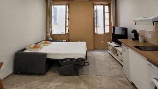 Une chambre d’hôtel de taille réduite, comprenant un lit, une petite kitchenette, un téléviseur et une fenêtre avec des rideaux.