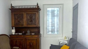 A living room with a grey sofa, a wooden cabinet, a wicker chair, and a window with blinds.