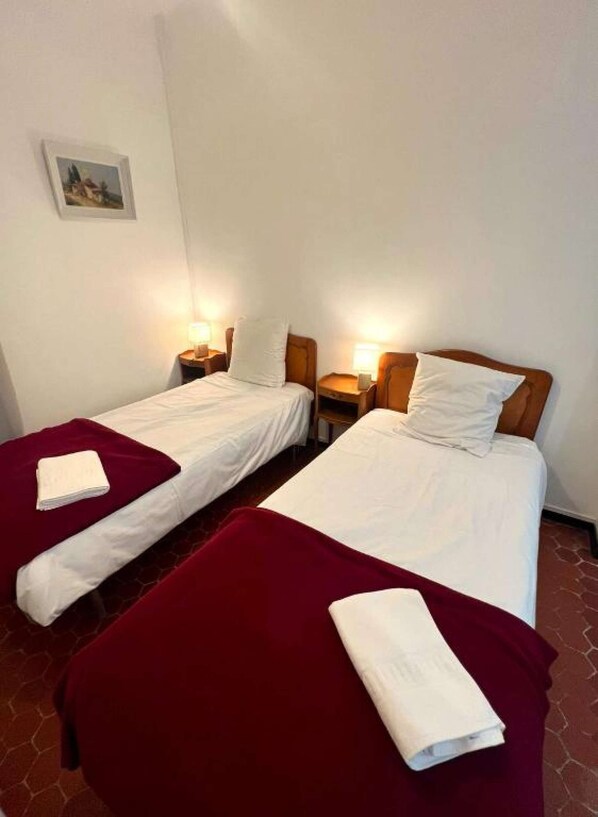 Deux lits simples avec des têtes de lit en bois, du linge de lit blanc et des plaids rouges. Chaque lit est muni d’une table de chevet avec une lampe.