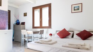 Habitación de hotel con cama, dos almohadas rojas, escritorio con sillas, una mesita con un florero y una ventana.
