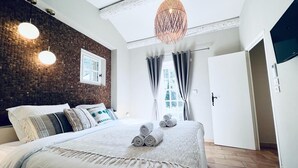 Et moderne soveværelse med en murstensvæg, en stor seng med sammenrullet håndklæder, en hængende lampe og en dør til et andet rum.
