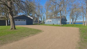 A gravel driveway leading to a house with a garage, surrounded by trees and a body of water in the background. - Sunset Harbor Retreat,3 bed, 2 bath w/ private launch and dock in charming Isle (Isle)