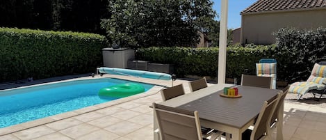 A swimming pool with a slide, a covered patio area with a table and chairs, and a hedge in the background.
