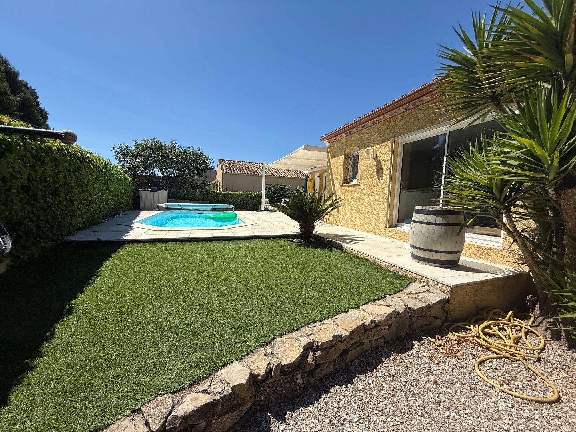 Un pati d'habitatge amb una piscina, gespa artificial, mur de retenció de pedra i una palmera.