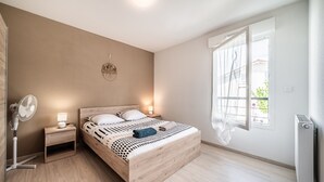 A bedroom with a wooden bed, bedside lamps, a standing fan, and a window with curtains. - Apartment "Le Cocon" with Mountain View, Balcony, and Wi-Fi (Thonon-les-Bains)