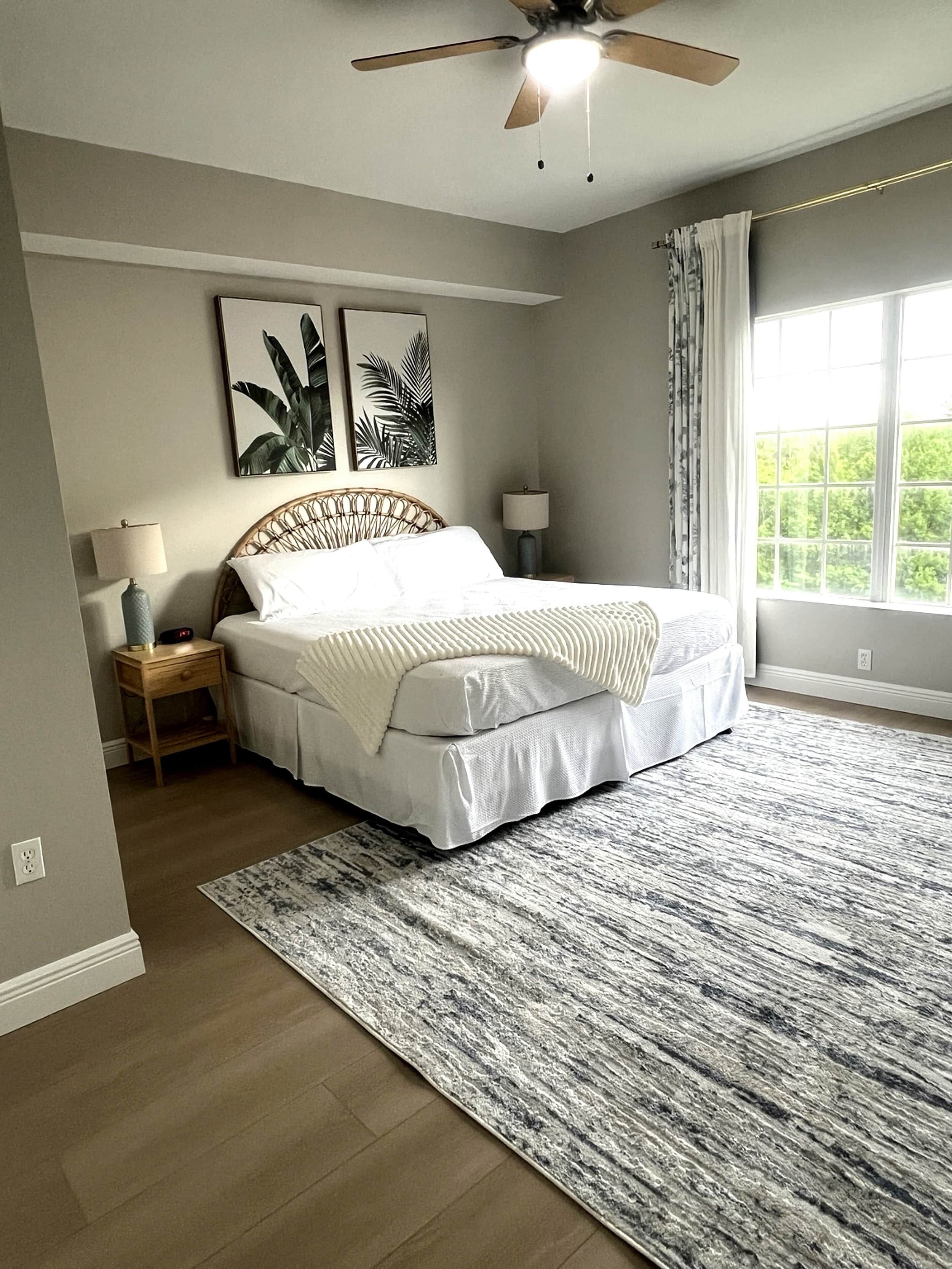 A bedroom with a bed, two bedside tables, a ceiling fan, a window with curtains, and a large rug.
