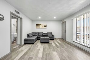 Ein modernes Wohnzimmer mit einem Ecksofa, einem kleinen Hocker, einem Gemälde an der Wand und einem Fenster mit Jalousien.
