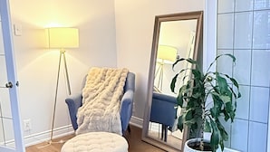 Ett rum med en rund pall, en blå fåtölj, en golvlampa, en spegel och en krukväxt.
