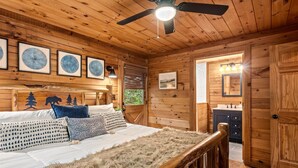 Interior de una cabaña de madera con cama, ventilador de techo, obras de arte enmarcadas y un baño con espejo y lavamanos.