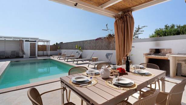 Zona de piscina exterior con mesa lista para comer, sillas y una parrilla.
