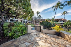 A stone-paved courtyard with palm trees, a gate, and a building with a red roof in the background. - NEW Kaanapali Royal Large Ground Floor 2-Bedroom Condo with Complimentary Rental Car with 6+ Nights! KRO-F102 by KBM (Lahaina)