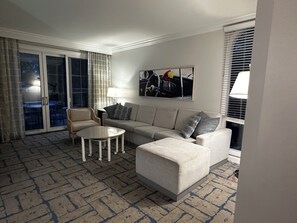 Ein Wohnzimmer mit Sofa, Sessel und Couchtisch, eine Essecke mit Tisch und Stühlen sowie ein großes Fenster mit Jalousien.