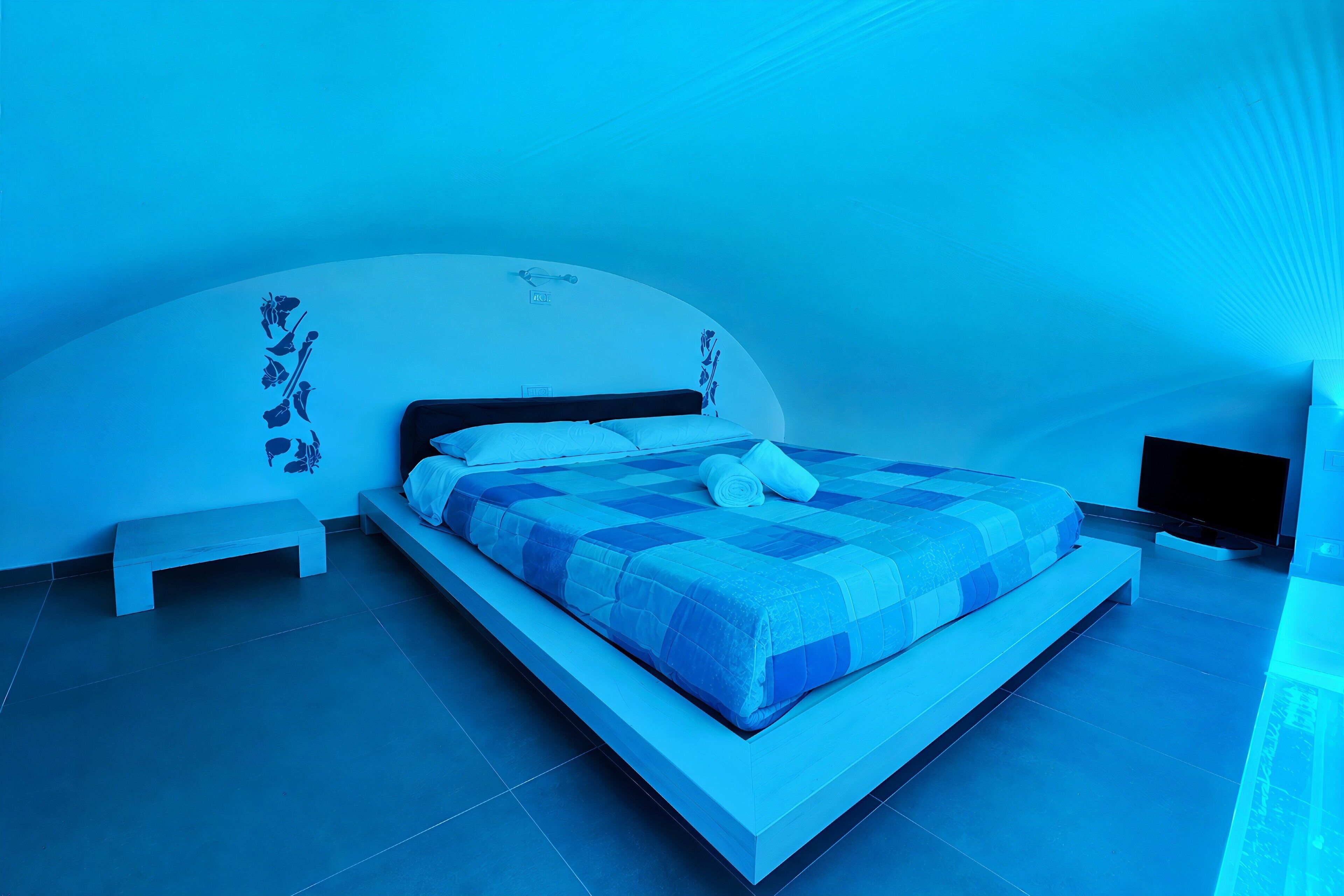 A modern bedroom with a large bed, a bench, and a flat-screen TV.