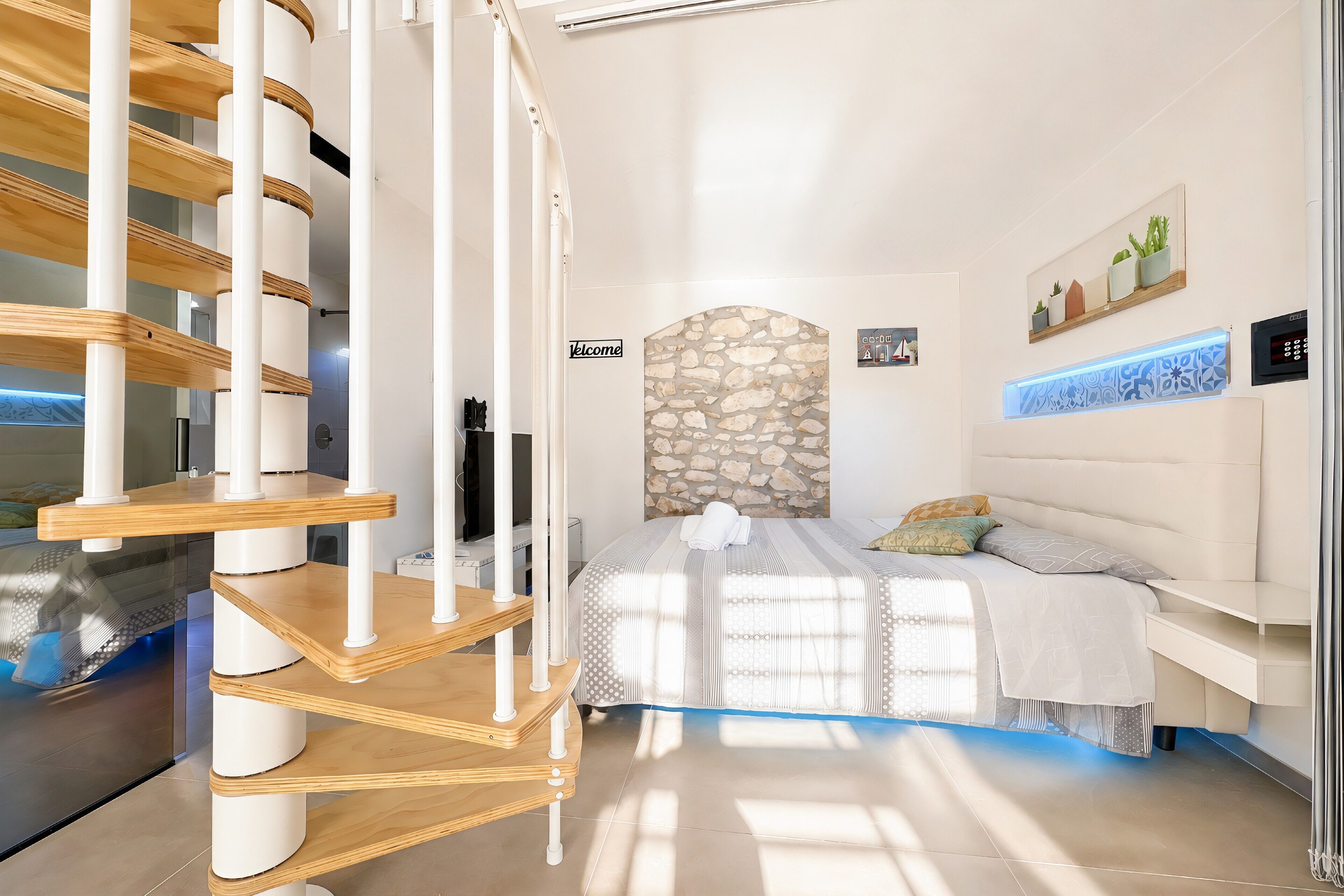 A modern bedroom with a spiral staircase, a stone accent wall, a bed with a striped bedspread, and a wall-mounted shelf with decorative items.