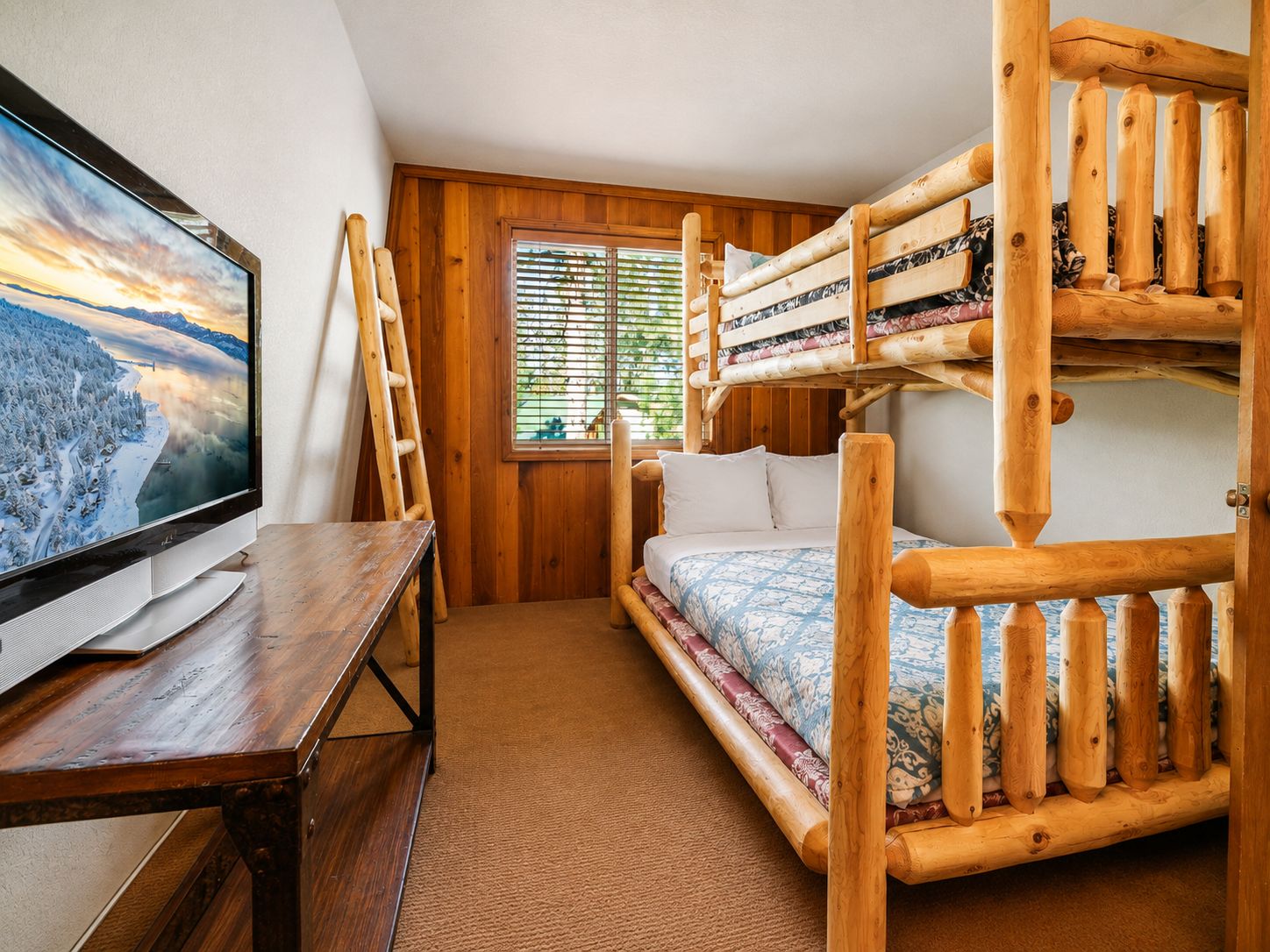 Une chambre avec un lit superposé, une télévision posée sur une table en bois, une fenêtre avec des stores et un sol recouvert de moquette.