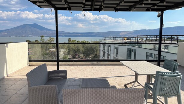 Een overdekt terras met rieten meubels, een tafel en stoelen, met uitzicht op een waterpartij en verre bergen.