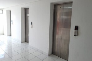 Un pasillo con tres puertas de ascensor, cada una con un panel de control. El suelo está cubierto de baldosas y las paredes son blancas.
